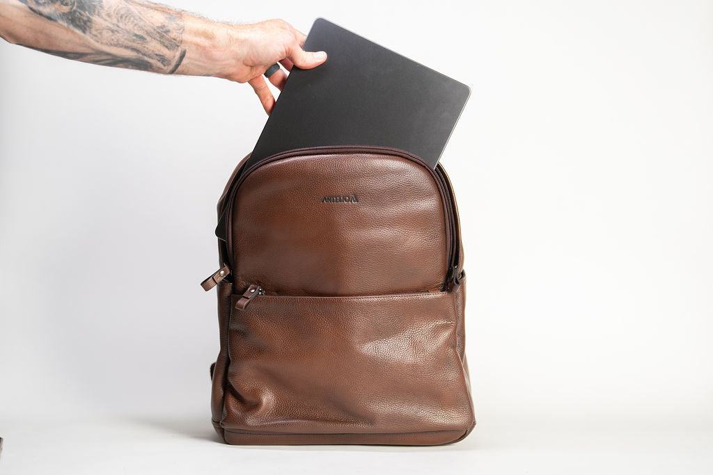 A laptop being placed inside the Artelio brown leather backpack to show the main compartment capacity