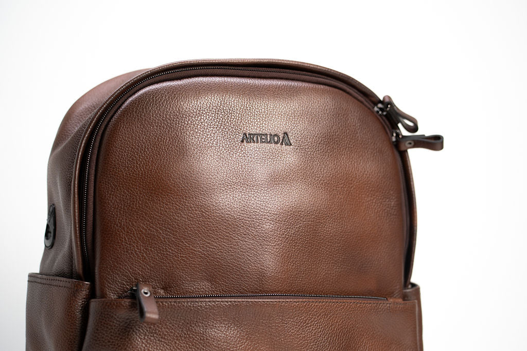 Upper front detail of the Artelio brown leather backpack showing the logo and curved zipper